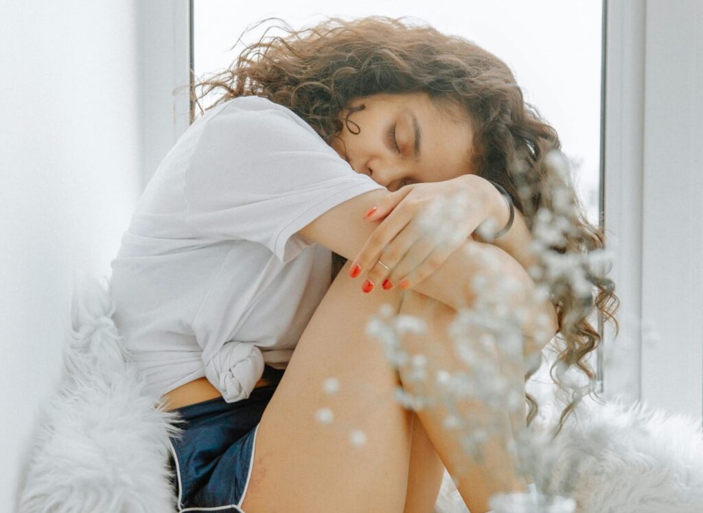 Person sitzt zusammengerollt und traurig auf einer Fensterbank und hat die Augen geschlossen.